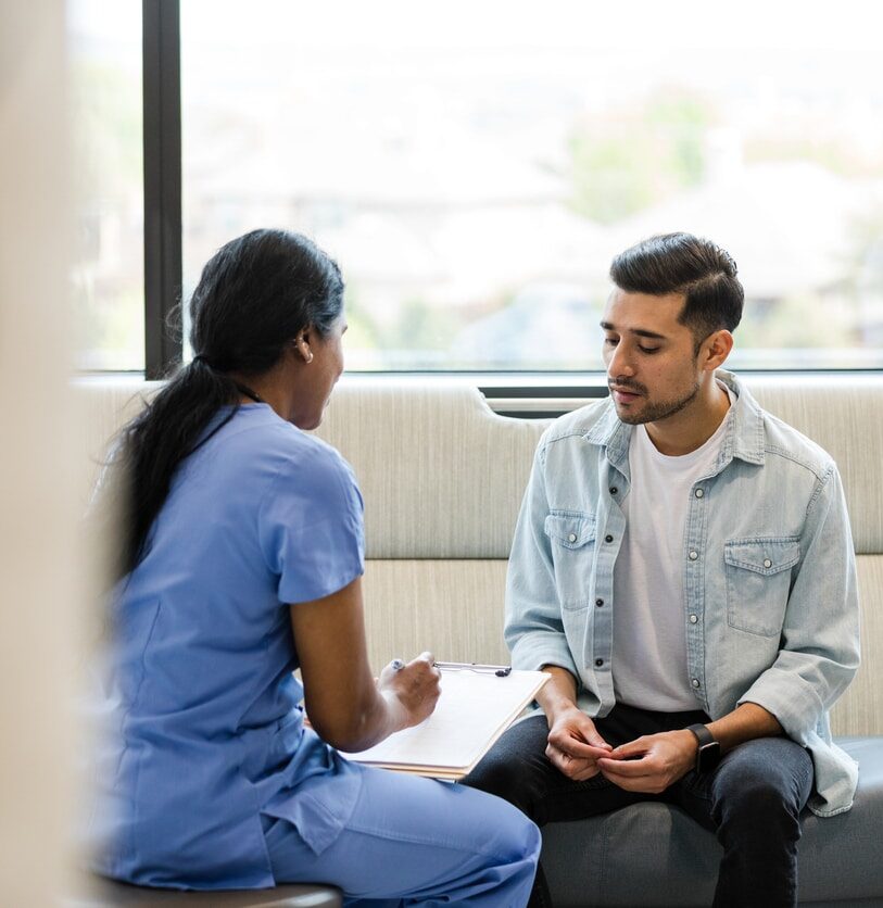 The young adult male sits on the hospital room couch to talk with the hospital insurance specialist to update medical information.