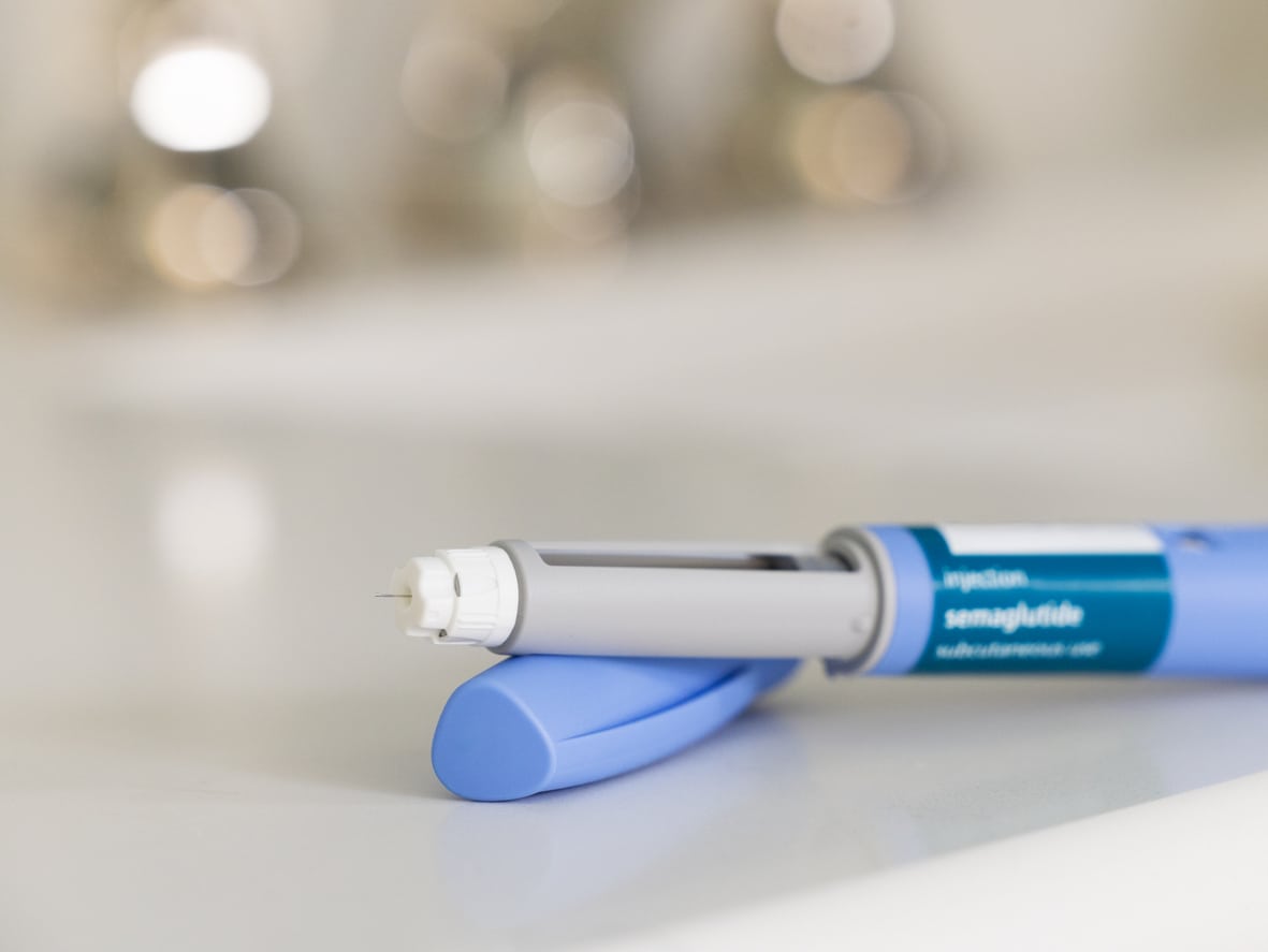 GLP-1 semaglutide injection pen, a medication used to treat Type 2 diabetes and aid in weight loss, on a white surface. The prescription medicine is used to treat Type 2 diabetes as It helps regulate blood sugar levels by stimulating insulin secretion and decreasing glucagon levels. In addition, it is also used off-label for weight loss in individuals with obesity, as it can help reduce appetite and increase feelings of fullness. The pen is positioned horizontally, with its cap removed, on a bathroom countertop with a white surface. The medical device is in the foreground and the background is out of focus because of a shallow depth of field.