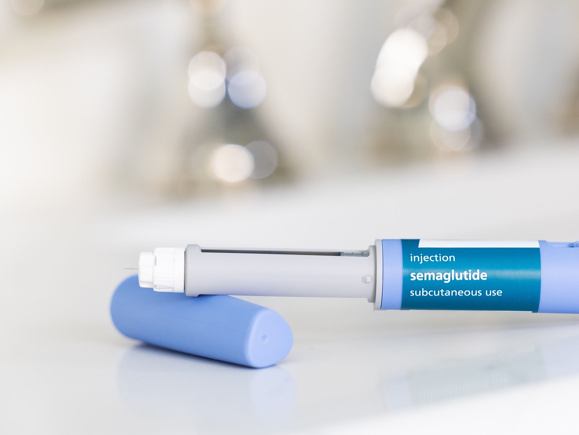 GLP-1 semaglutide injection pen, a medication used to treat Type 2 diabetes and aid in weight loss, on a white surface. The prescription medicine is used to treat Type 2 diabetes as It helps regulate blood sugar levels by stimulating insulin secretion and decreasing glucagon levels. In addition, it is also used off-label for weight loss in individuals with obesity, as it can help reduce appetite and increase feelings of fullness. The pen is positioned horizontally, with its cap removed, on a bathroom countertop with a white surface. The medical device is in the foreground and the background is out of focus because of a shallow depth of field.